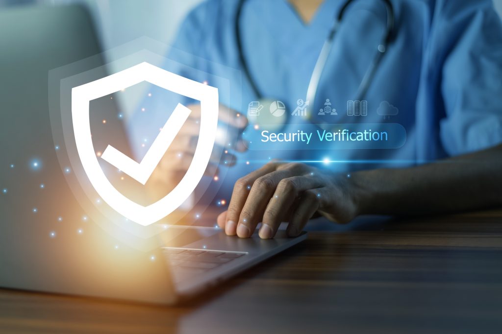 A person in medical scrubs types on a laptop, with a large digital shield and checkmark icon overlay and the words "Security Verification" symbolizing secure access to healthcare data.