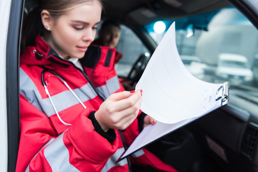 A paramedic wearing a red and gray jacket sits in an emergency vehicle, holding and reviewing a clipboard with paperwork. A stethoscope hangs around her neck, and another person is partially visible in the background.