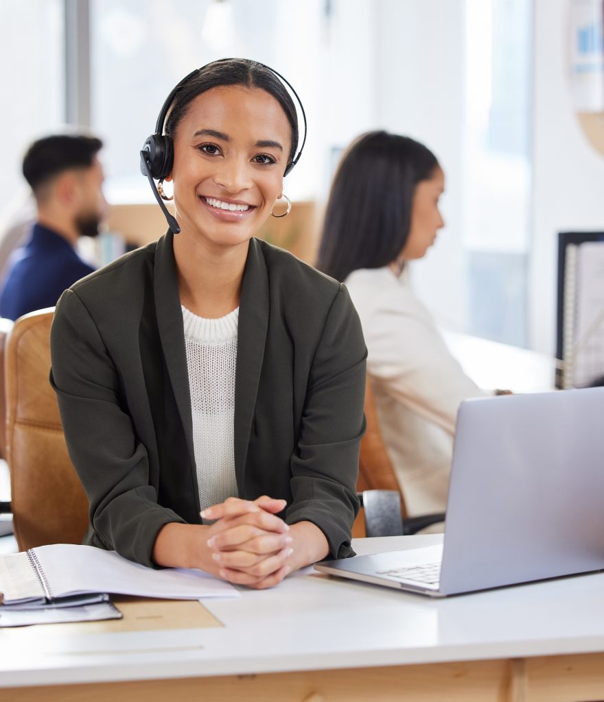A smiling woman wearing a headset sits at a desk with a laptop, notepad, and pen. She faces the camera, while two colleagues work in the background in a bright office setting.